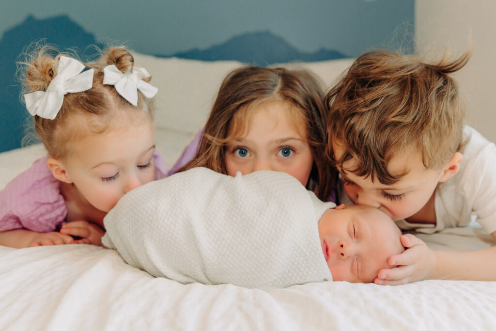Asheville Newborn Photographer captures three young children - two girls and one boy - are lying stomach down on a bed with white covers, gently kissing their newborn sibling. The newborn is wrapped in a white cloth newborn wrap expertly wrapped by the asheville newborn photographer.