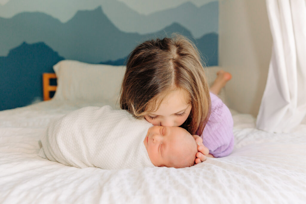 Big Sister kisses newborn baby brother on the head while lying on a bed during in-home lifestyle newborn session in Asheville, North Carolina with Newborn Photographer Rachel Marie Photography