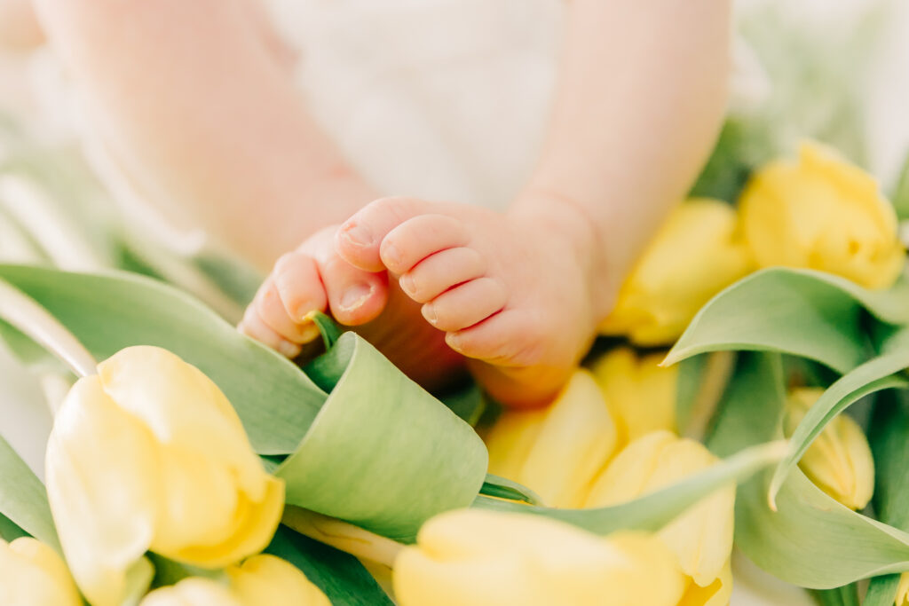 Asheville Newborn Photographer captures a close-up of a baby's feet resting on a bed of fresh yellow tulips with green leaves, capturing a soft and tender moment in a bright, natural setting.