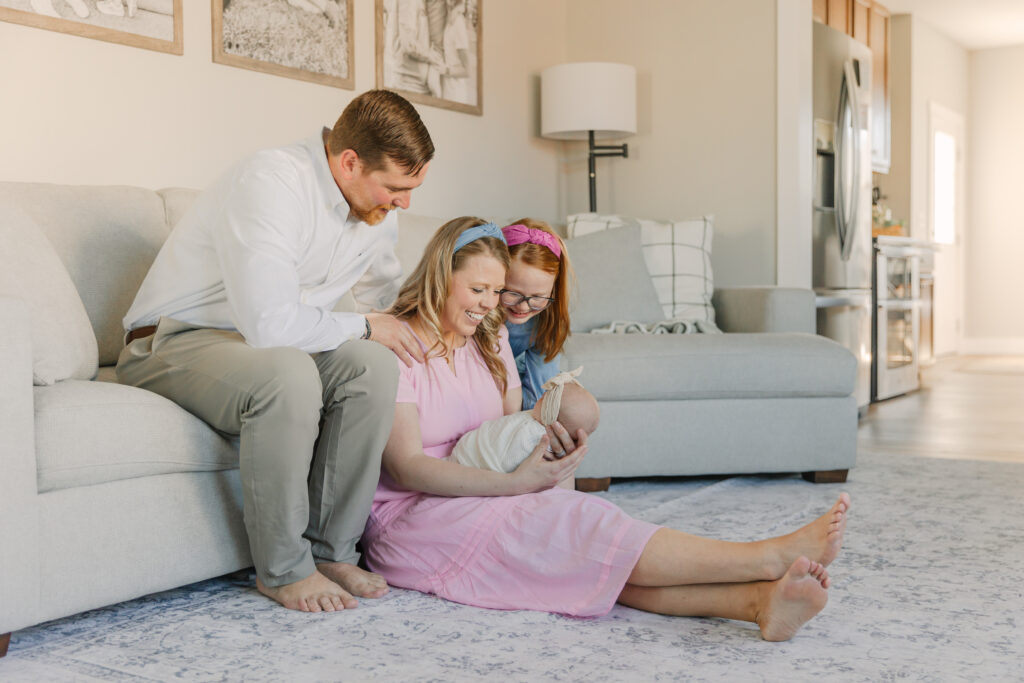Asheville Newborn Photographer captures a joyful family sitting together in a cozy living room. A postpartum mother in a pink dress sits on the floor holding a newborn baby, smiling warmly. A father sits beside her on the couch, and a new big sister with red hair and a pink headband leans in close, all admiring the baby with love and tenderness.