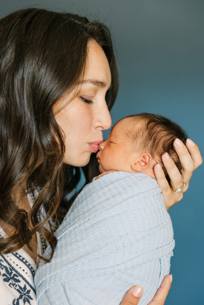 Asheville Newborn Photographer captures a tender close-up of a new mother gently kissing her newborn baby’s nose. The baby is wrapped snugly in a soft blue newborn wrap, resting peacefully against the mother's chest. The mother holds the baby lovingly with both hands, her eyes closed in a moment of deep affection.