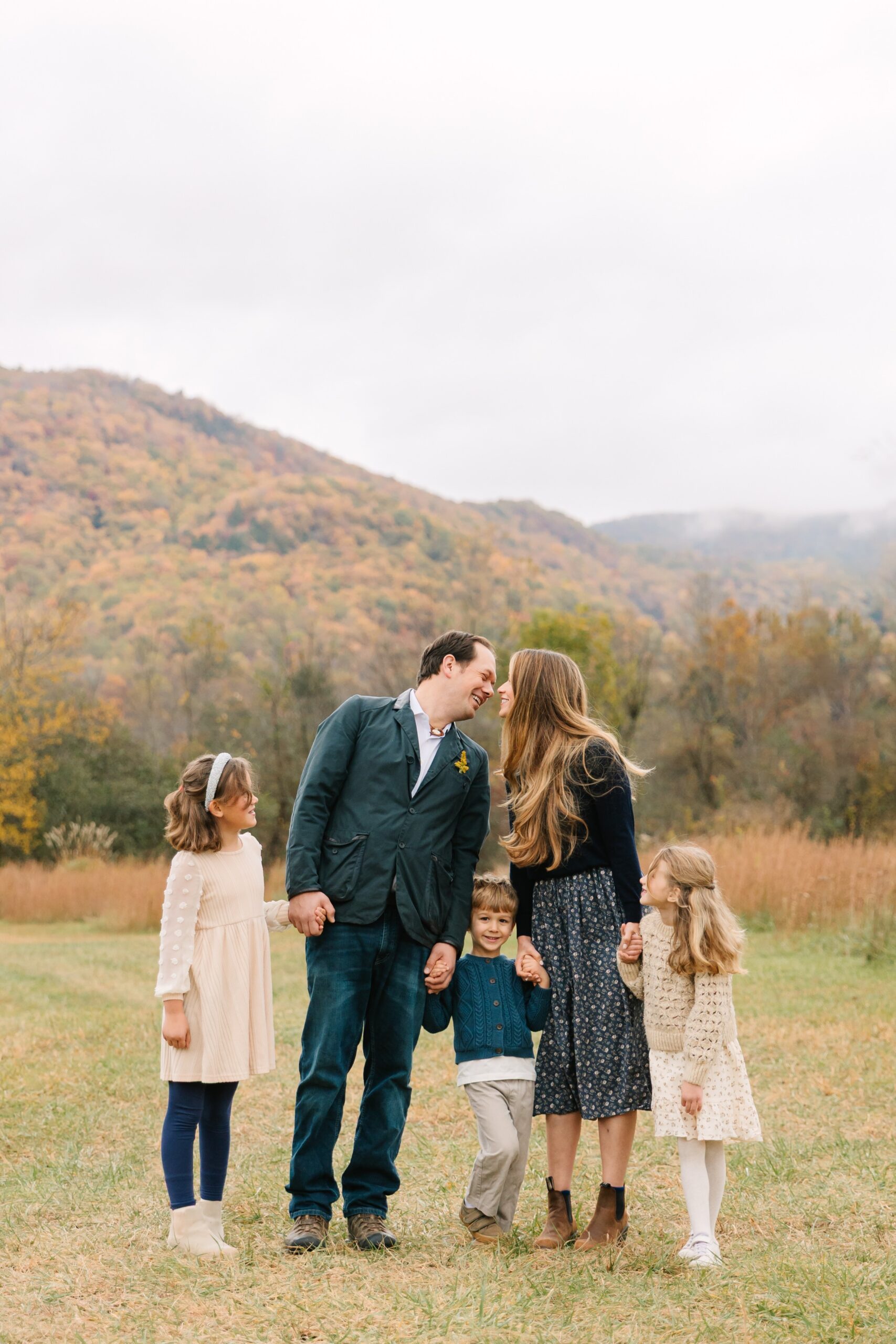 Family with three young kids holds hands and mom and dad lean in for a kiss during fall family photo session in front of mountain views with asheville family photographer rachel marie photography
