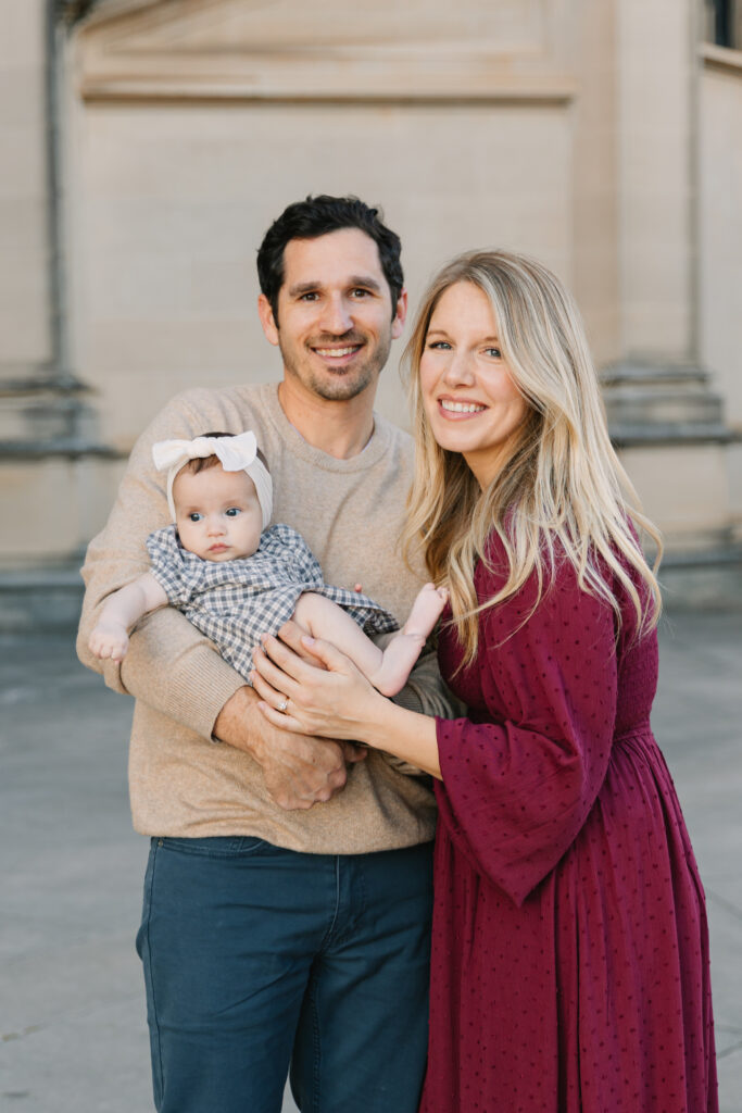 Family of three smiling in front of Biltmore House in a photo taken by Asheville Family Photographer. Mom is smiling while touching baby girl's leg and dad is holding baby girl.