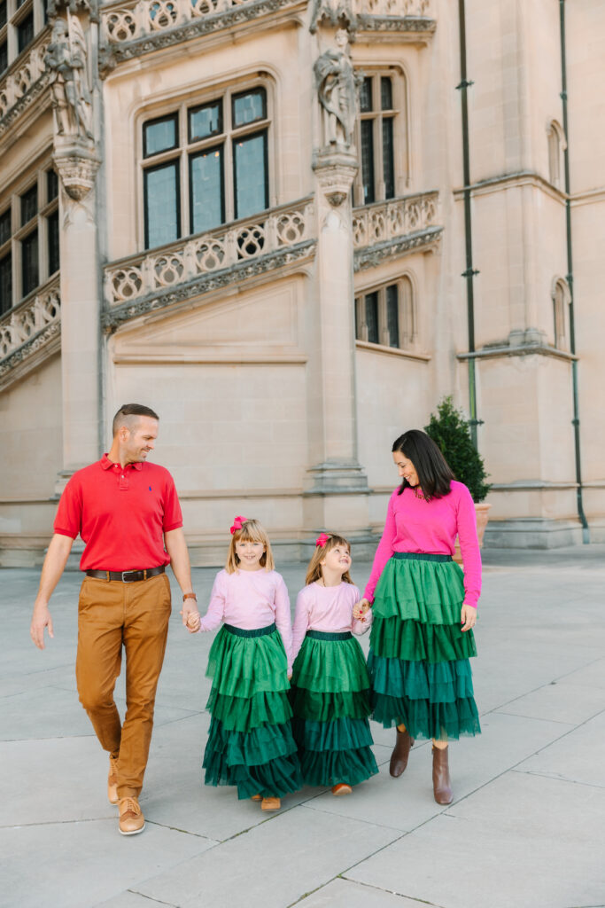 Family of four pictured walking and laughing while holding hands in front of the Biltmore House.