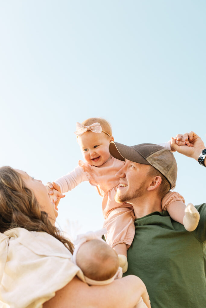 Family show smiling at one another and having fun. Toddler is on dad's shoulders and smiling down at mom and newborn baby sister. 