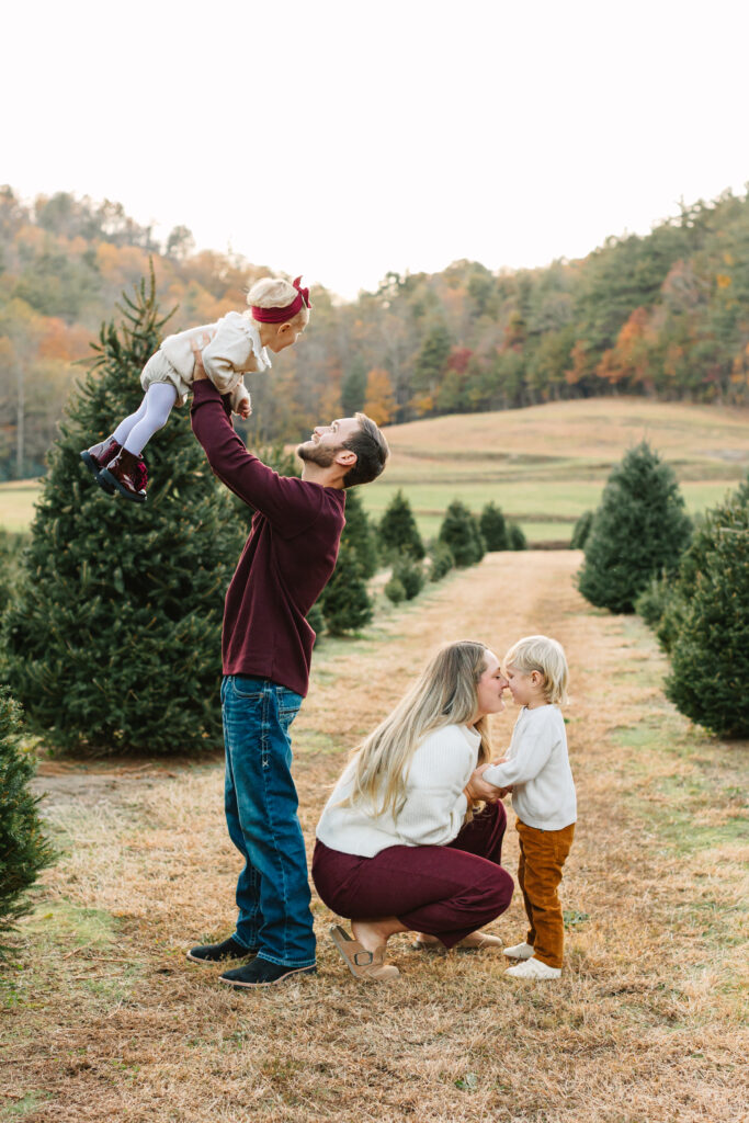 Family of four posing for photo at a Christmas tree farm. Mom is crouched and giving her son a kiss while dad is smiling and tossing his daughter in the air.