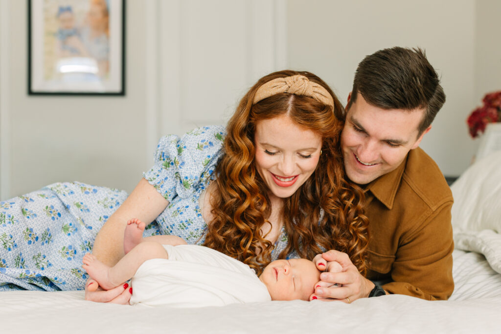 Mom and dad lay on bed and smile down at newborn baby boy during in-home newborn session with Asheville Newborn Photographer