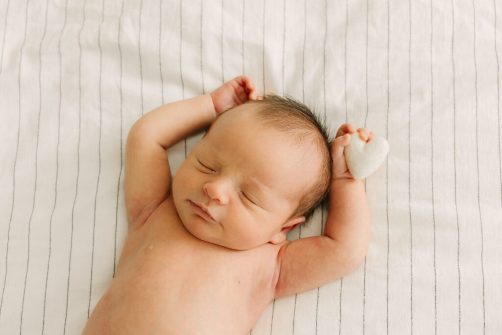 newborn baby boy lies on white bedding with arms stretched high and grasping onto a little white cotton heart during in-home newborn photo session in Asheville, North Carolina with Rachel Marie Photography