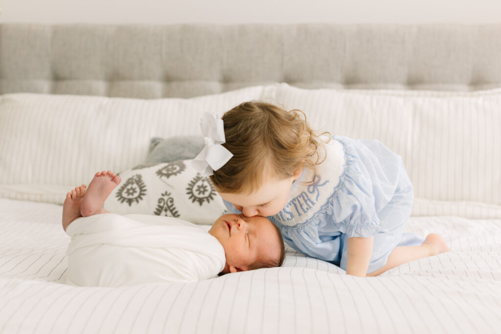 Toddler sister kisses newborn baby brother on bed during Asheville In-Home Newborn Photography Session