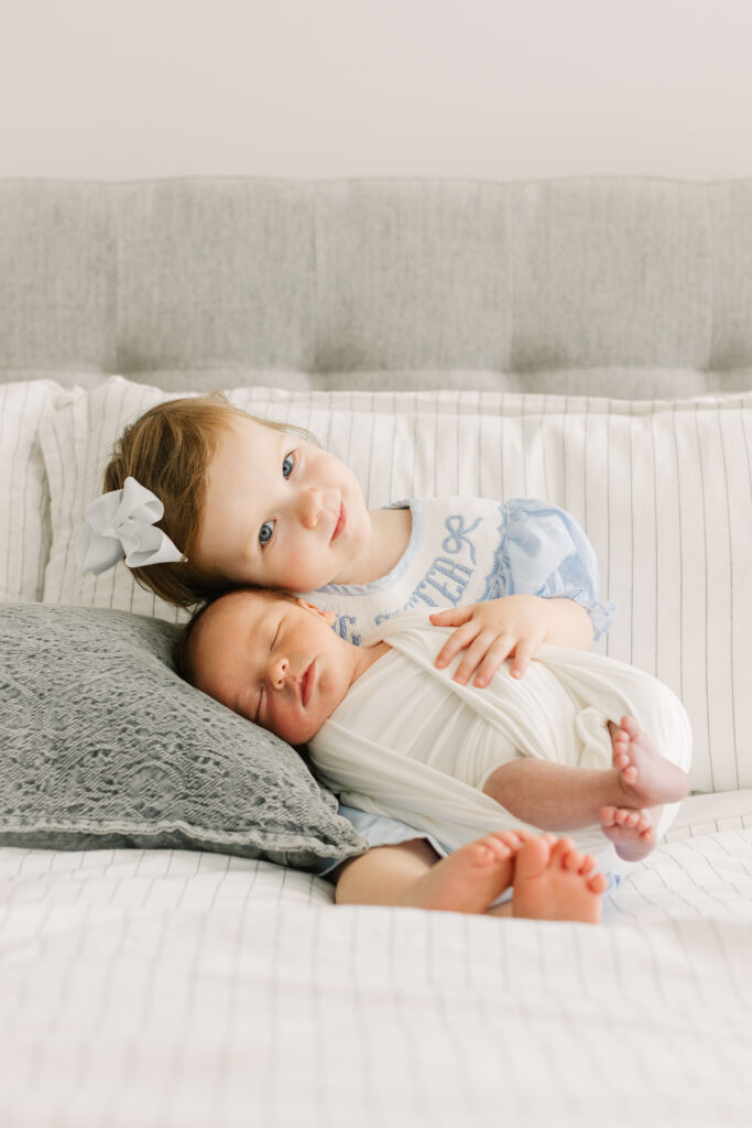 big sister holding newborn brother on bed. baby is in a while swaddle and big sister has a blue dress and white bow on during Asheville in-home newborn session