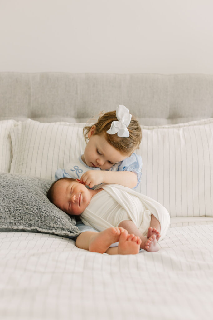 big sister holding newborn brother on bed. baby is in a while swaddle and big sister has a blue dress and white bow on 