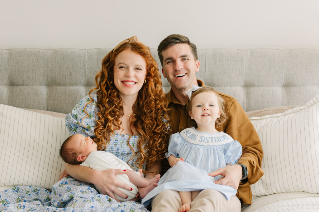 Family of four pictured smiling on their bed for a photo. Mom is holding their newborn son and dad is holding their young daughter. Family is dressed in white, blue, and tan colors