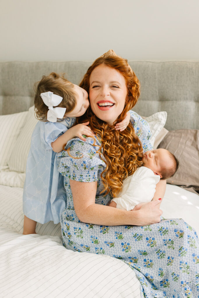Mom of two in blue floral hill house dress holding her newborn son while her toddler daughter kisses her on the cheek during in-home newborn session with Asheville Newborn Photographer Rachel Marie Photography
