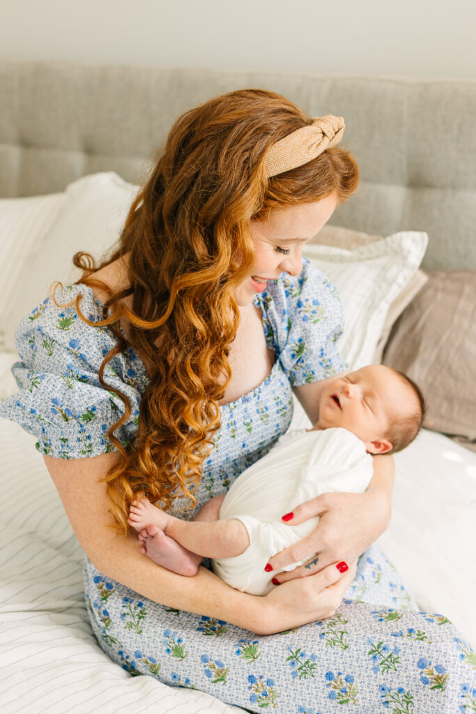 mom in blue floral dress holding her newborn son on the side of the bed during in-home newborn session with Asheville Newborn Photographer Rachel Marie Photography