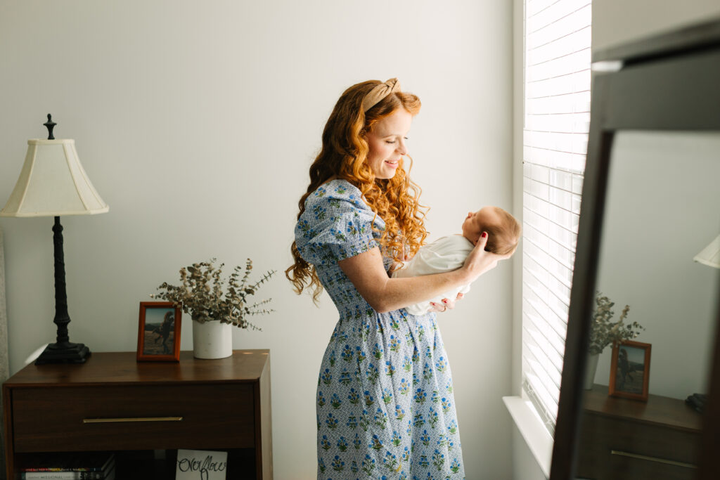mom in blue floral dress holding her newborn son in front of window
