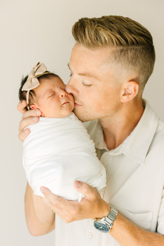 Dad photographed kissing newborn daughter on the cheek. Newborn is swaddled in a white swaddle and is wearing a pink bow.