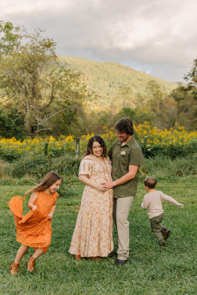 Maternity photo session in a field with family of 4. Mom and dad are embracing and smiling at other children while the two children are running around mom and dad.