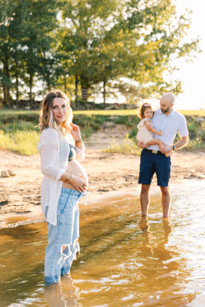 Family posing for maternity photos while wading in the edge of a lake. Mom is smiling and holding her belly while dad and daughter are in the background embracing. 