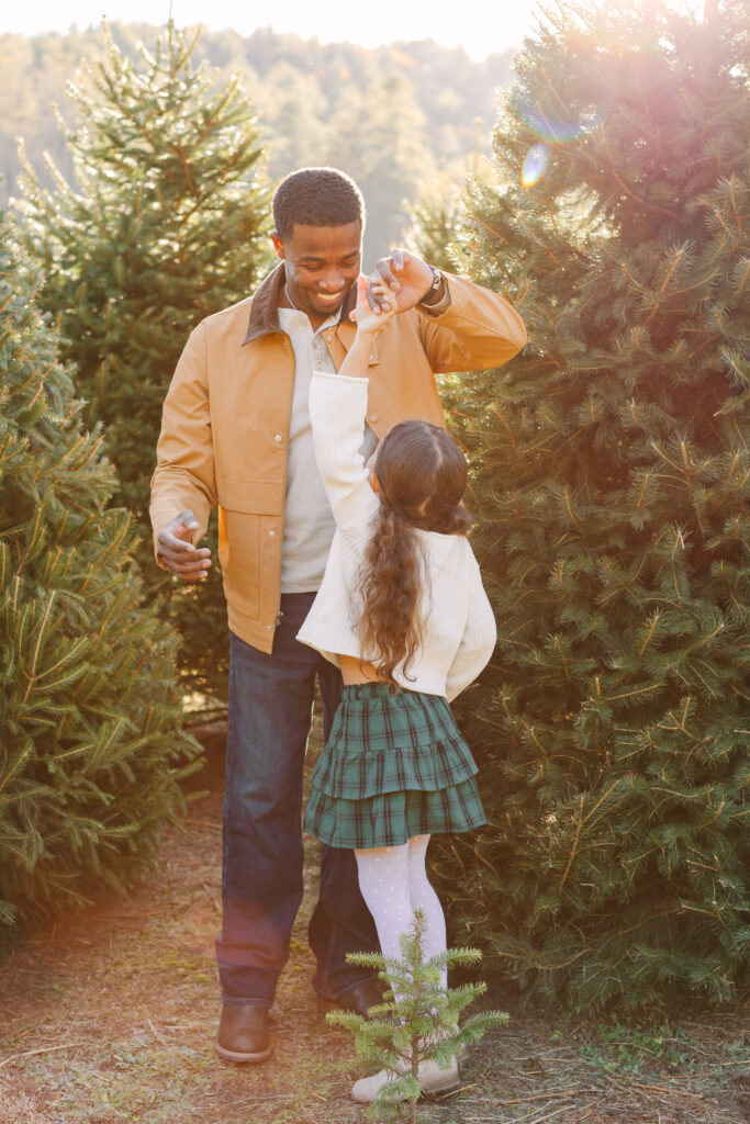 Dad pictured spinning/ dancing with daughter at a Christmas tree farm.