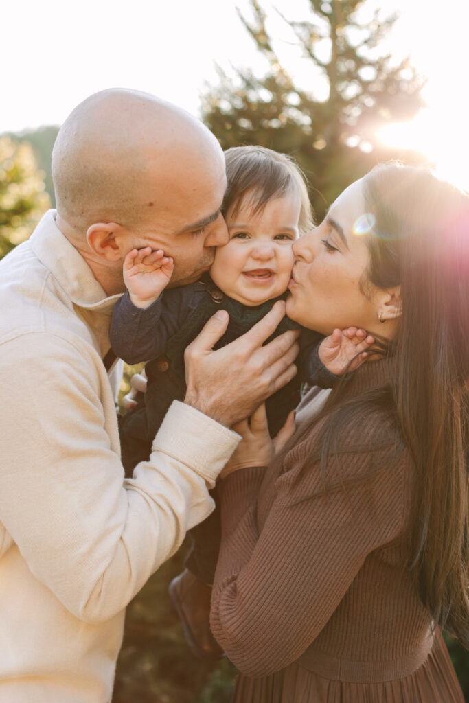 Mom and dad kissing son on each cheek at a Christmas tree farm.