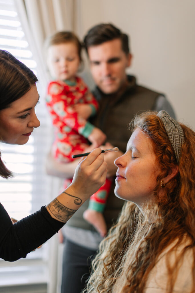 Postpartum mom getting make-up applied by professional stylist for her in-home newborn session while her husband and toddler watch from behind