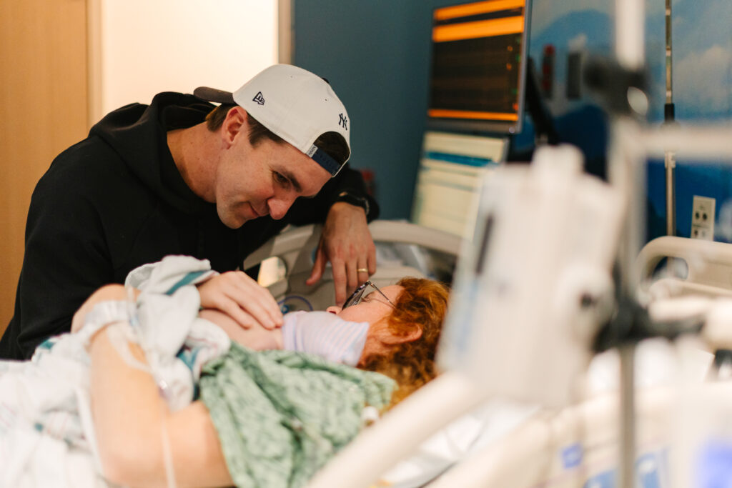 mom lays in hospital bed with newborn baby boy on her chest while dad leans over to look at him just after delivery at mission hospital in Asheville, NC