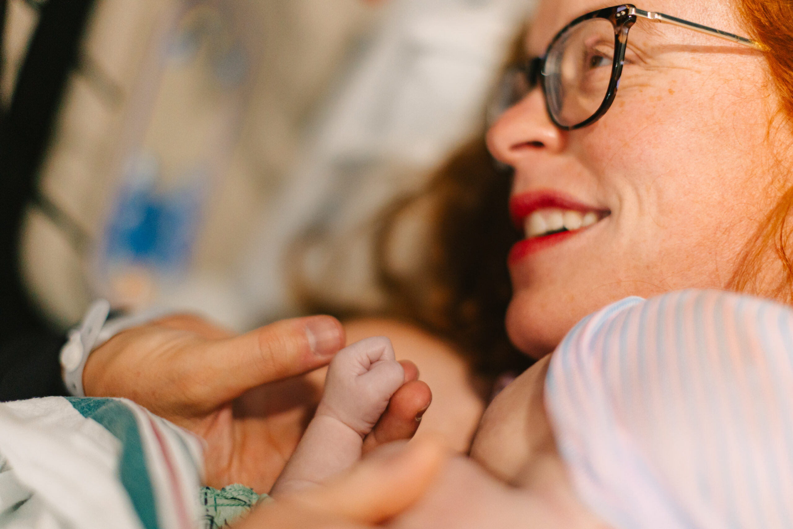mom delivers birth to baby son and smiles with him laying on her chest in hospital bed while dad reaches down to hold his tiny hand during Asheville birth photography session at Mission Hospital with Rachel Marie Photography