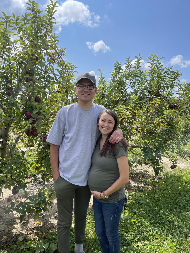 pregnant couple smiles at an apple orchard in Hendersonville during asheville babymoon