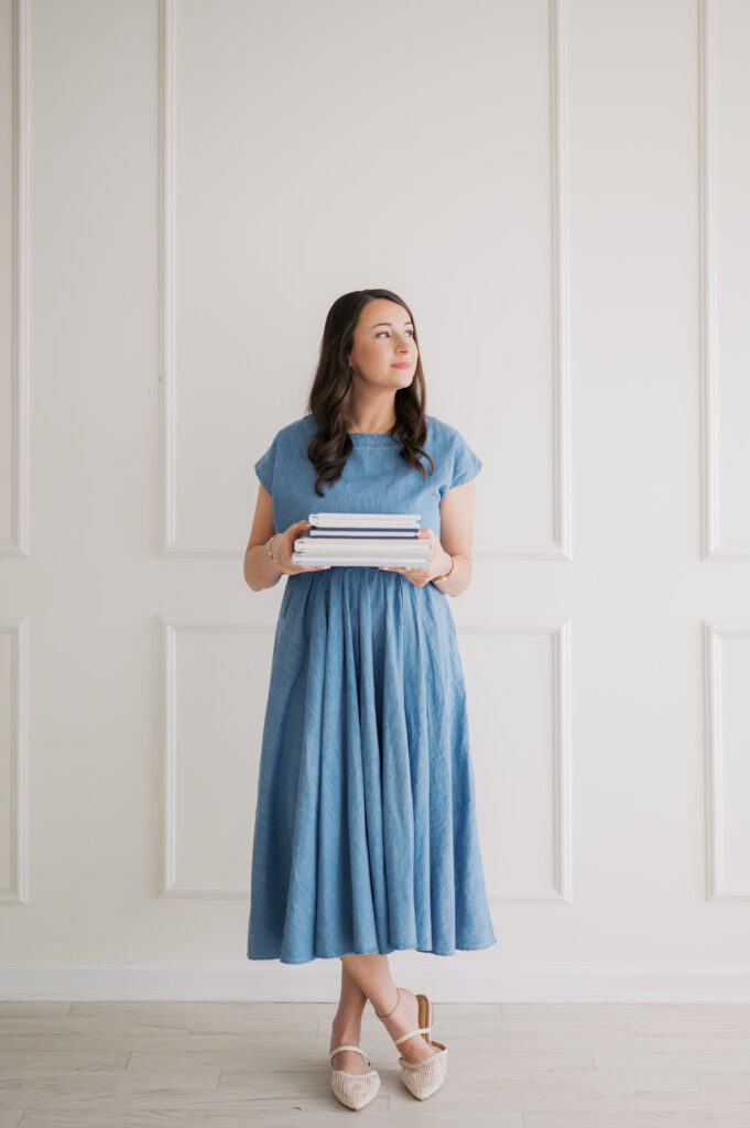 Photographer stands in blue dress and holds several photo albums to showcase the print products she provides for her family photography clients in the asheville, nc area