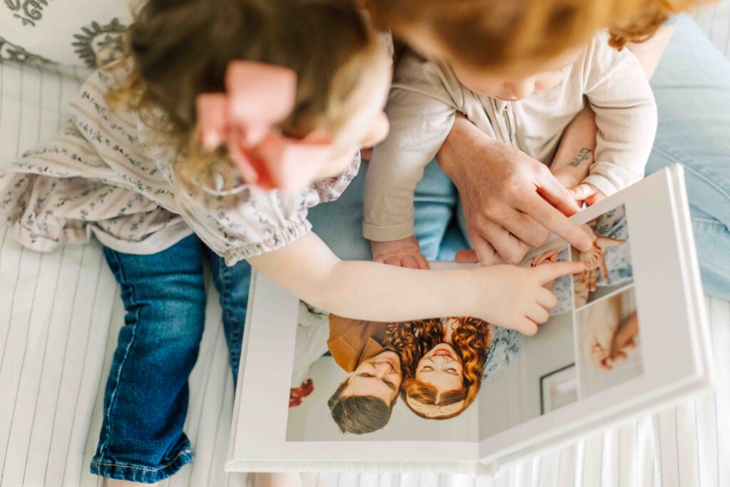mom sits on bed with her children and looks through the RMP Signature Photo Album that their photographer designed for them with photos from their newborn session in Asheville, NC