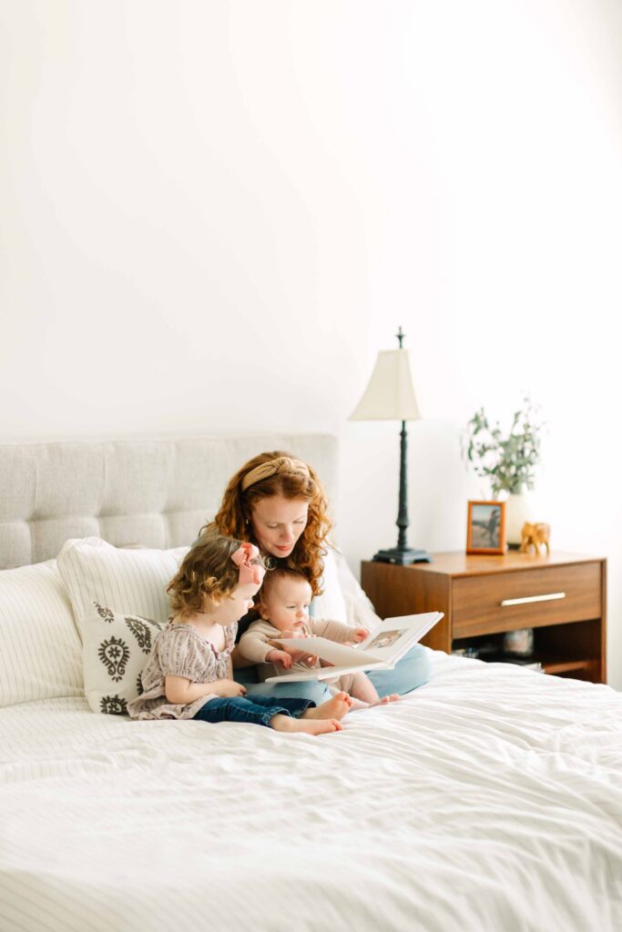 Mom sits on the bed with her toddler daughter and baby son and looks at a family photo album with their family's pregnancy announcement, birth and newborn photos in it.