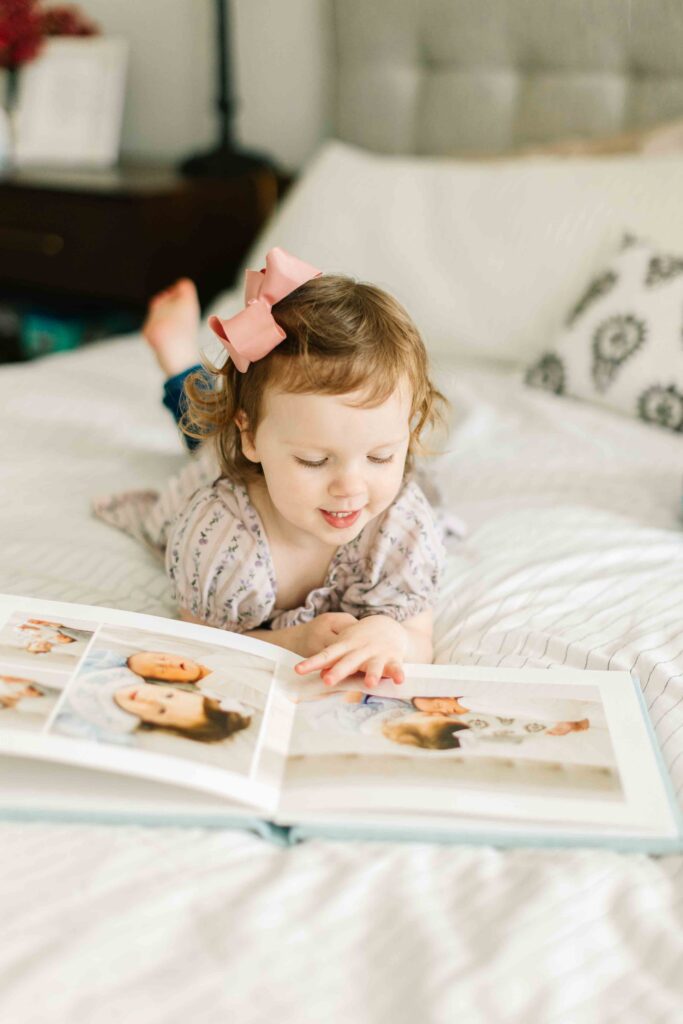 toddler girl lays on bed and looks through newborn photo session in her family photo album designed by asheville newborn photographer rachel marie photography