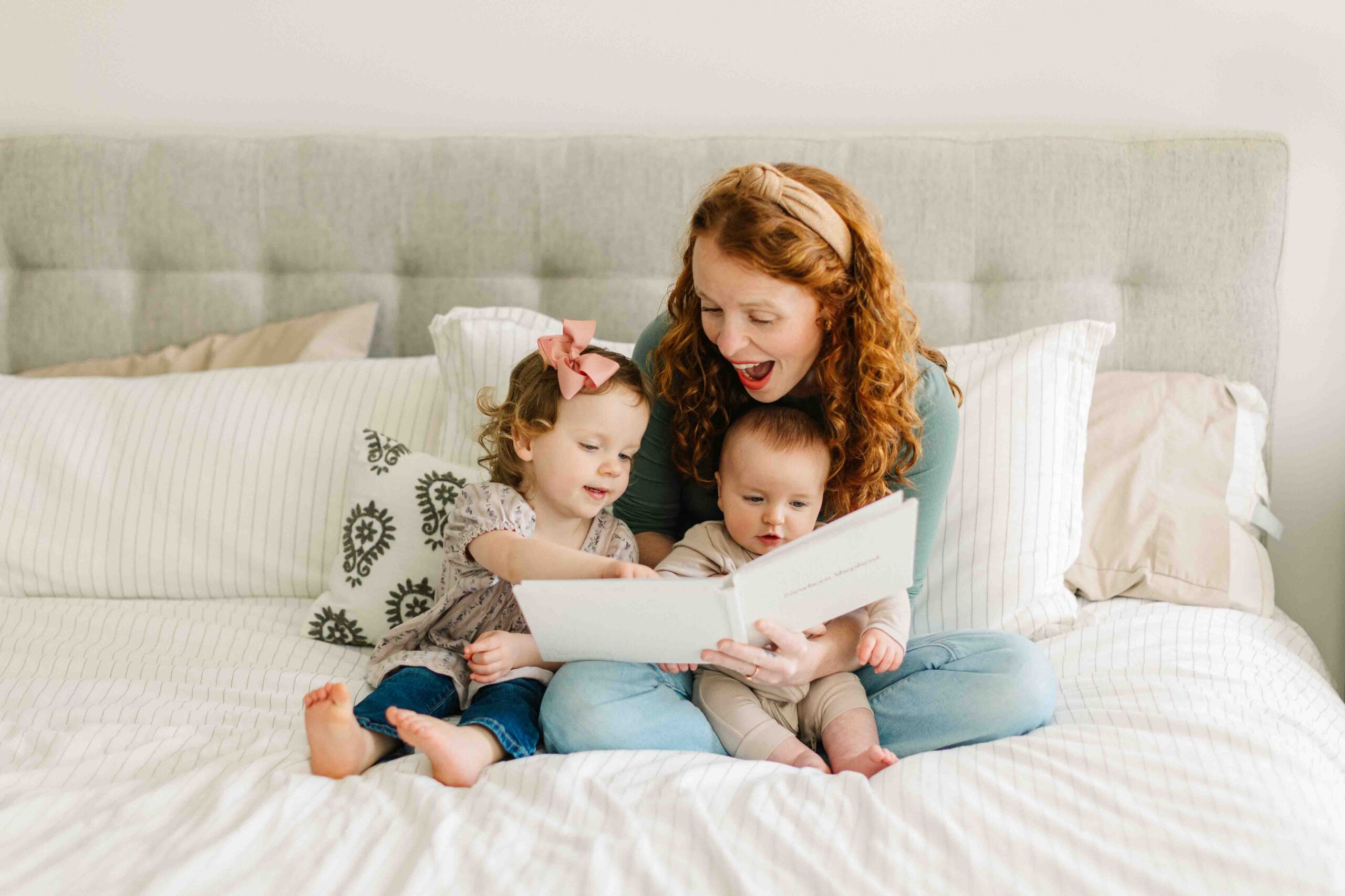 Mom sits on the bed with her toddler daughter and baby son and looks at a family photo album with their family's pregnancy announcement, birth and newborn photos in it.