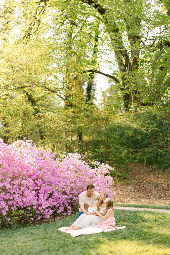 family smiles down at newborn baby girl while sitting on a blanket at the biltmore estate azalea newborn photo session in asheville, nc with asheville family photographer Rachel Marie Photography