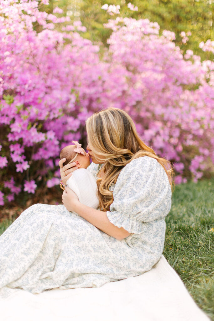 mom kisses newborn baby girl while sitting on a blanket in front of a large pink azalea bush at the Biltmore Estate Azalea Gardens in Asheville, NC
