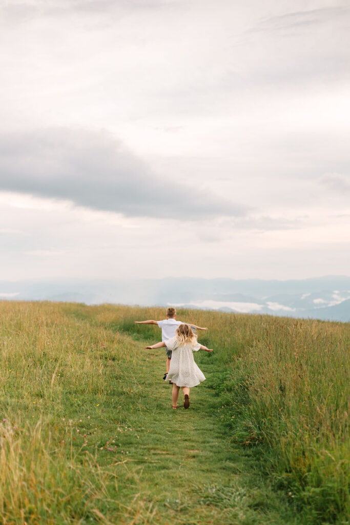 A brother and sister run through a field with airplane arms on top of Max Patch, NC near Asheville, NC during family photo session with Rachel Marie Photography